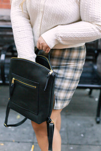 A woman wearing a cardigan and plaid skirt opens her black leather fashion backpack with gold zippers.

