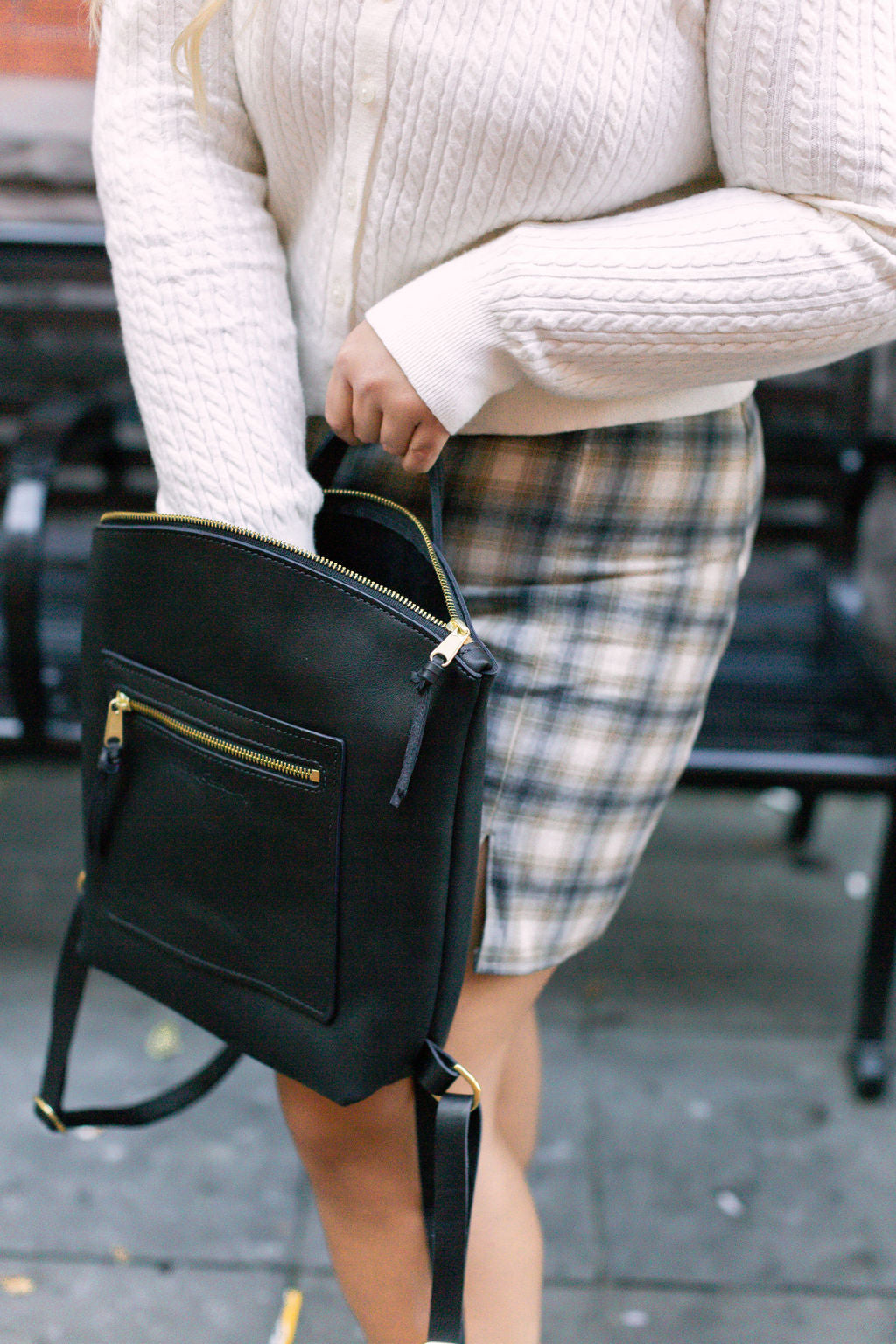 A woman wearing a cardigan and plaid skirt opens her black leather fashion backpack with gold zippers.
