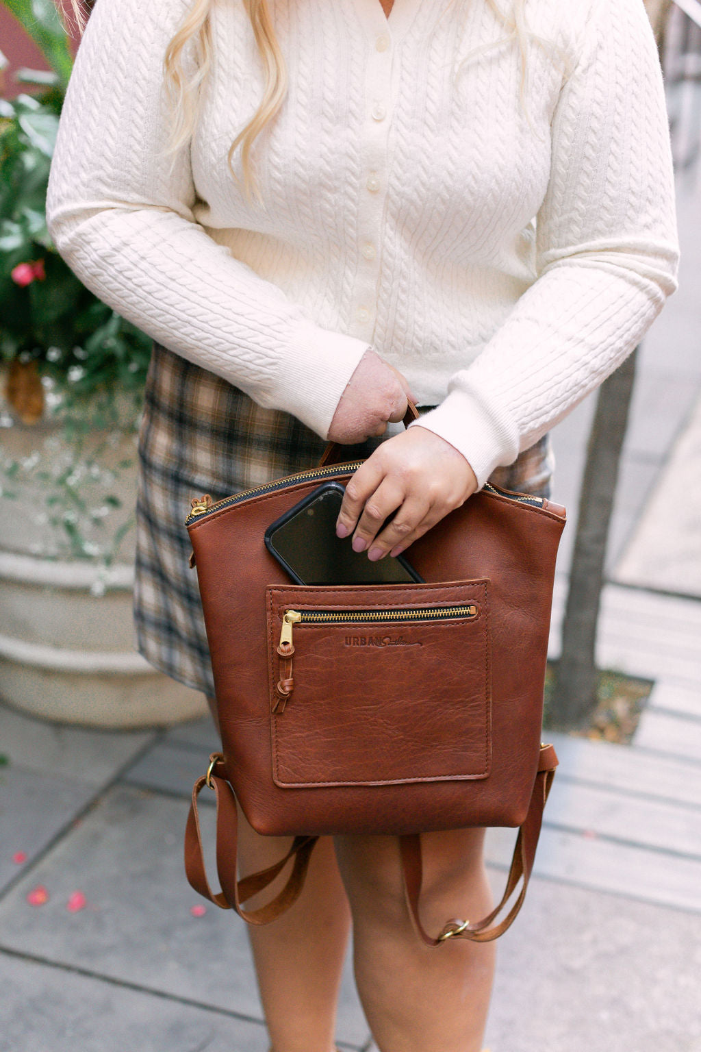 A woman places her smartphone into a brown leather Urban Originals backpack.
