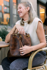 A blonde woman smiles while holding a stylish Urbane brown leather bucket bag at an outdoor cafe.
