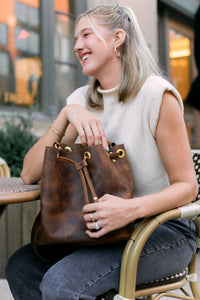 A blonde woman smiles while holding a stylish Urbane brown leather bucket bag at an outdoor cafe.
