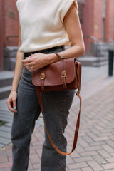 A woman models a stylish brown leather satchel handbag with brass buckles, paired with a cream sweater vest and grey jeans on a brick pathway.
