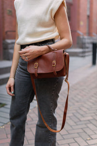 A woman models a stylish brown leather satchel handbag with brass buckles, paired with a cream sweater vest and grey jeans on a brick pathway.
