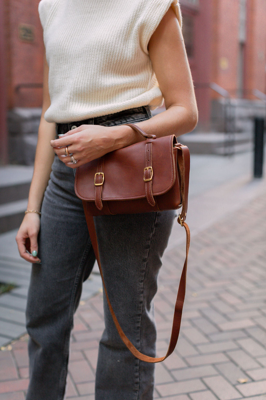 A woman models a stylish brown leather satchel handbag with brass buckles, paired with a cream sweater vest and grey jeans on a brick pathway.
