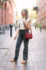 A blonde woman models a fashionable outfit with a red satchel bag while walking on a brick sidewalk.
