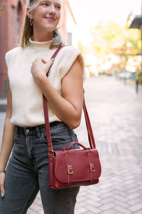 A stylish woman models a handcrafted burgundy leather satchel crossbody bag on a sunlit brick street.
