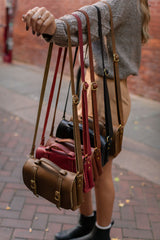 A woman displays a colorful assortment of handcrafted leather satchel handbags with adjustable straps.
