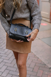 Woman carries a stylish black leather satchel purse with gold buckles, complementing her taupe sweater and tan skirt.
