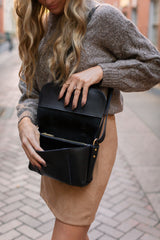 Woman opens a stylish black leather crossbody bag worn with a brown sweater and skirt on a city street.
