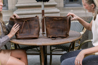 Two women admire handcrafted leather tote bags displayed on a cafe table.
