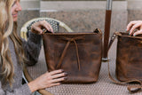 A woman showcases a rustic, brown leather bucket bag with a drawstring closure at an outdoor cafe.
