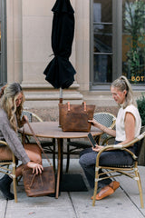 Two women display leather tote bags and a crossbody purse while sitting at a café table.
