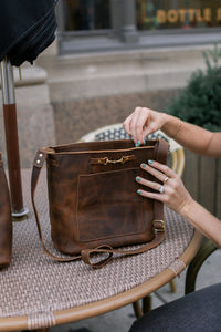 A woman reaches into a stylish brown leather crossbody bag on a wicker table.
