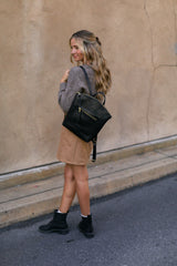 Woman walks wearing a stylish black leather backpack, tan skirt, grey sweater, and black boots.

