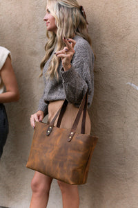 A woman models a stylish brown leather tote bag while wearing a gray sweater and suede skirt.
