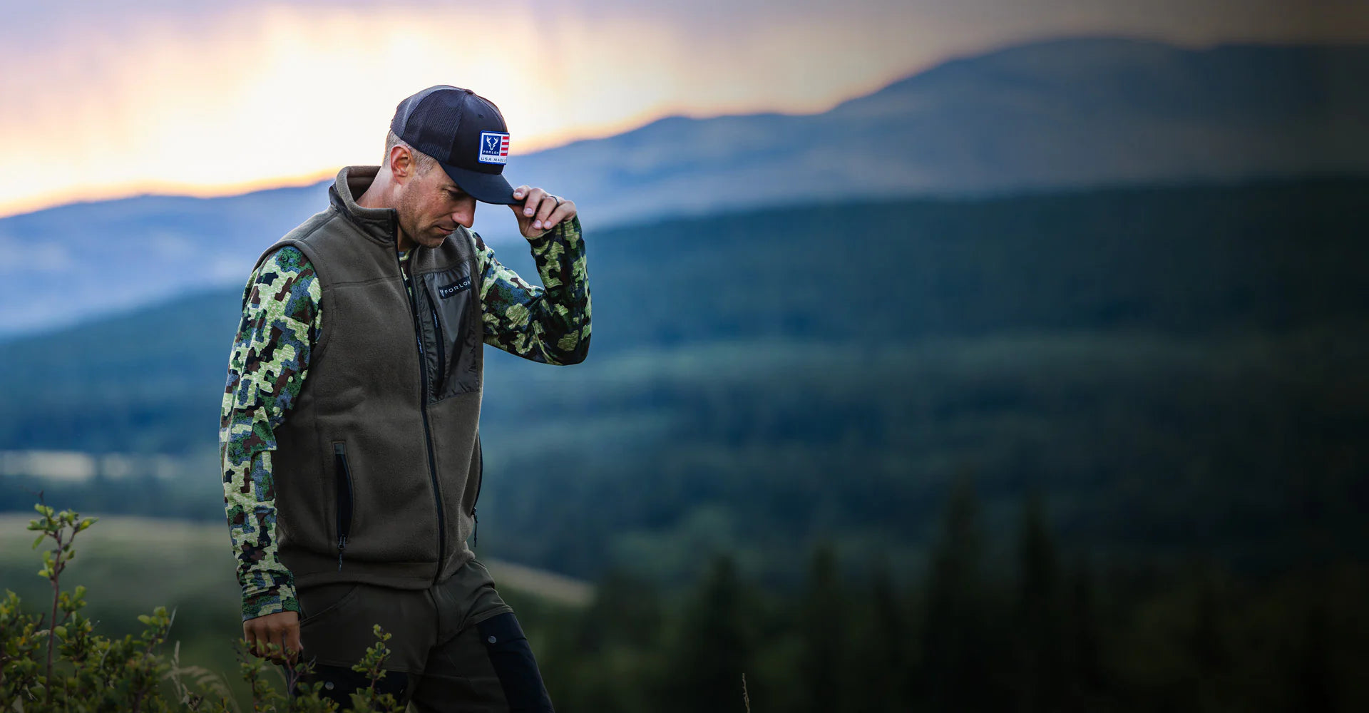 Man in Forloh camouflage shirt and vest and hat standing in a mountainous landscape with a sunset.