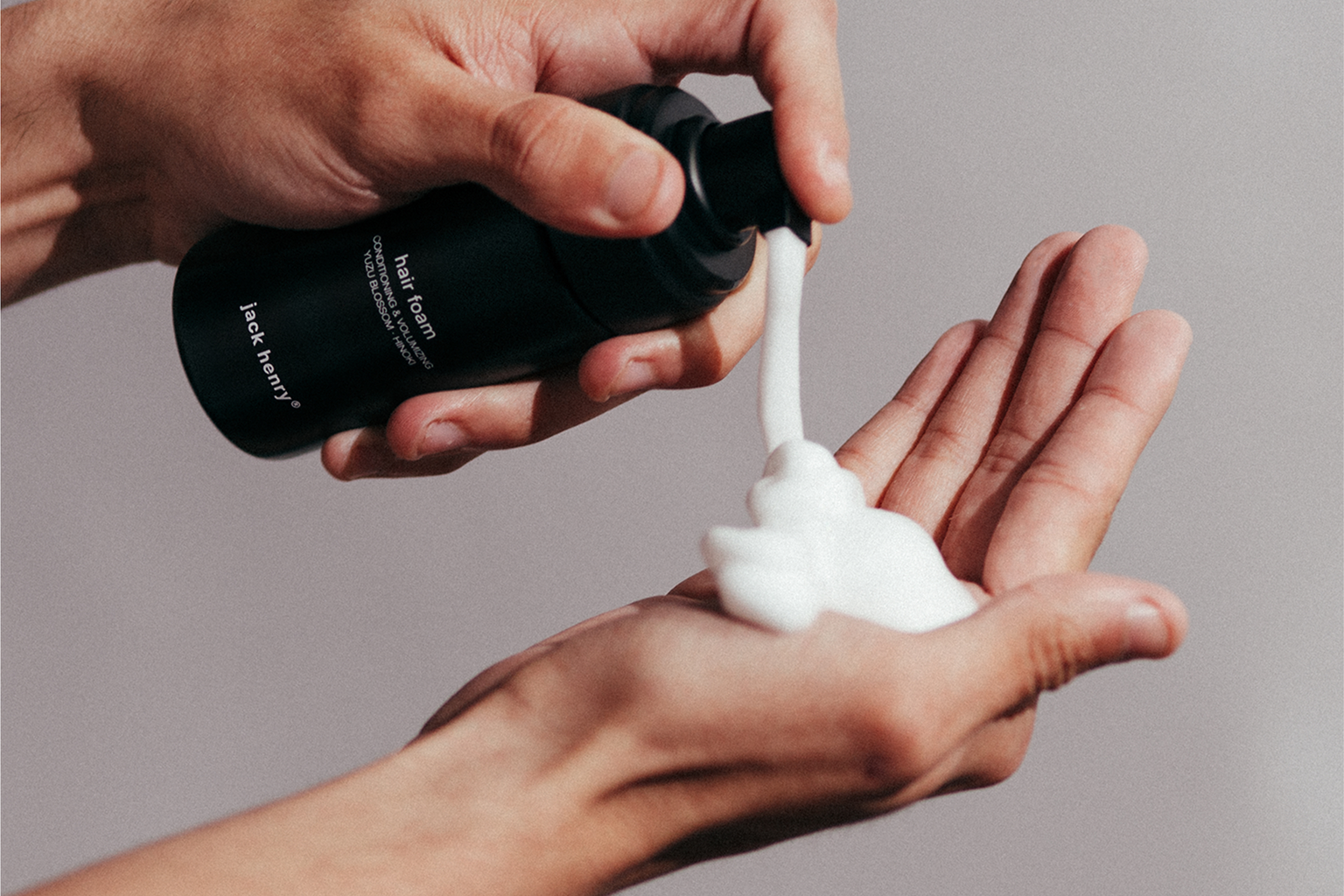 Person holding a black bottle and applying white cream to their hand against a neutral background