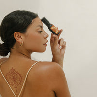 A woman with a back tattoo holds a bottle of green tea serum against her forehead.
