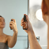 Woman sprays lavender and milky oat face mist in a bathroom mirror.
