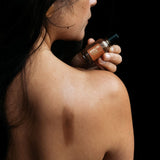 A woman displays her bronzed skin while holding a bottle of bronzing serum on her shoulder.
