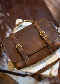 Artisan displays a handcrafted brown leather satchel with brass buckles atop a woven stool.
