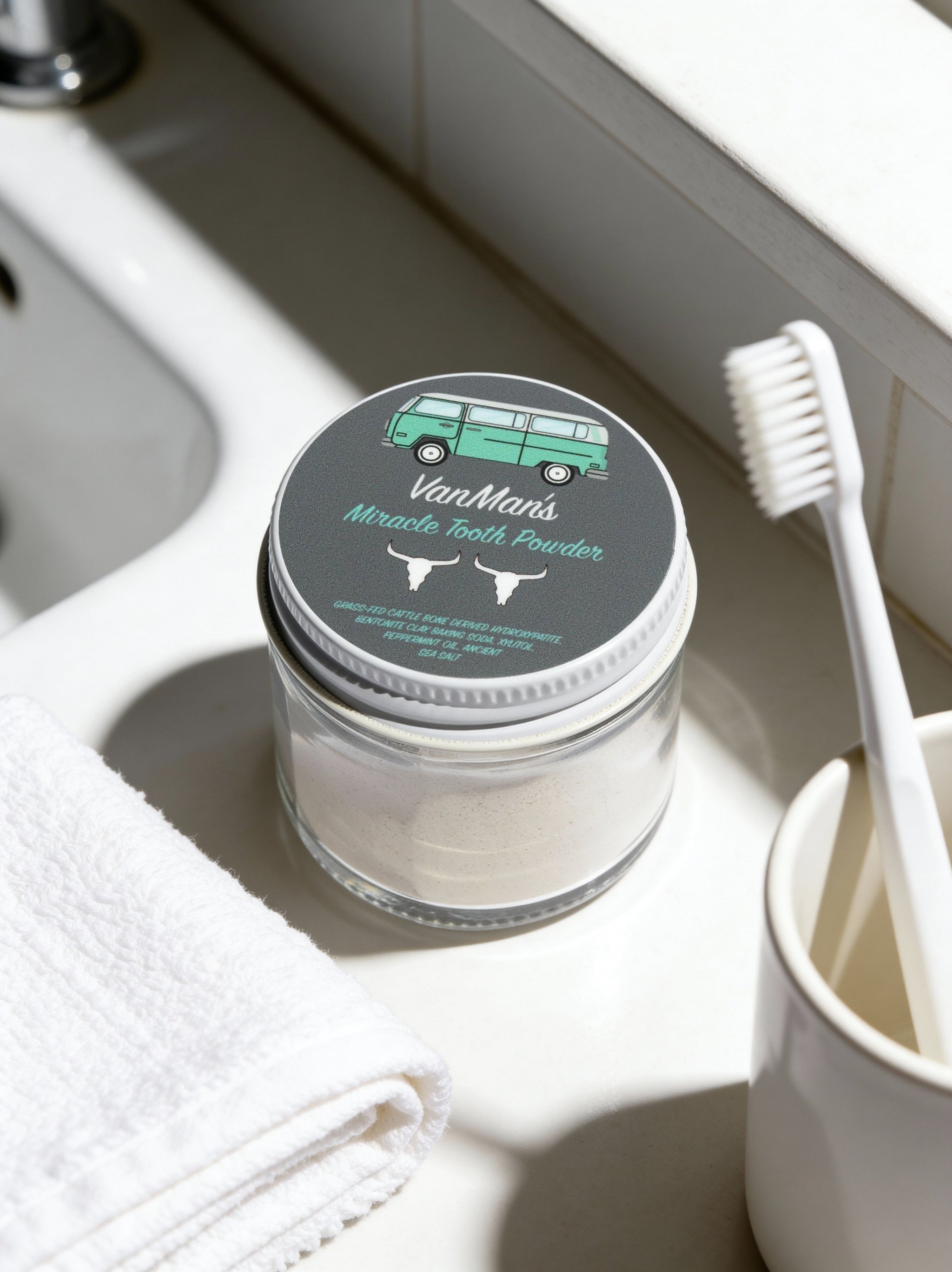 Jar of VanMan's Miracle Tooth Powder on a bathroom counter with a toothbrush and towel.