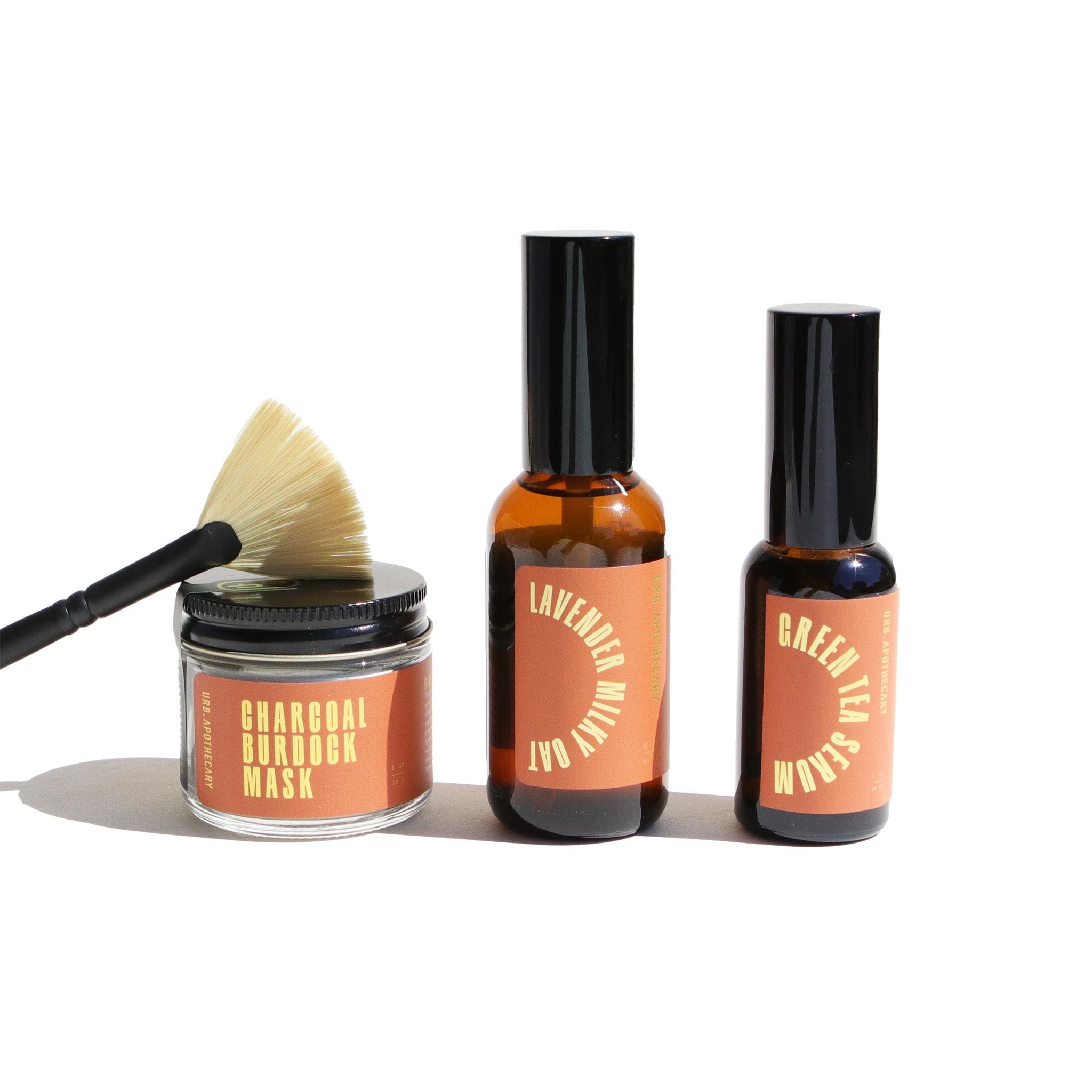 Arrange URB Apothecary skincare products, including a charcoal burdock mask, lavender milky oat lotion, and green tea serum, with a brush displayed against a white background.
