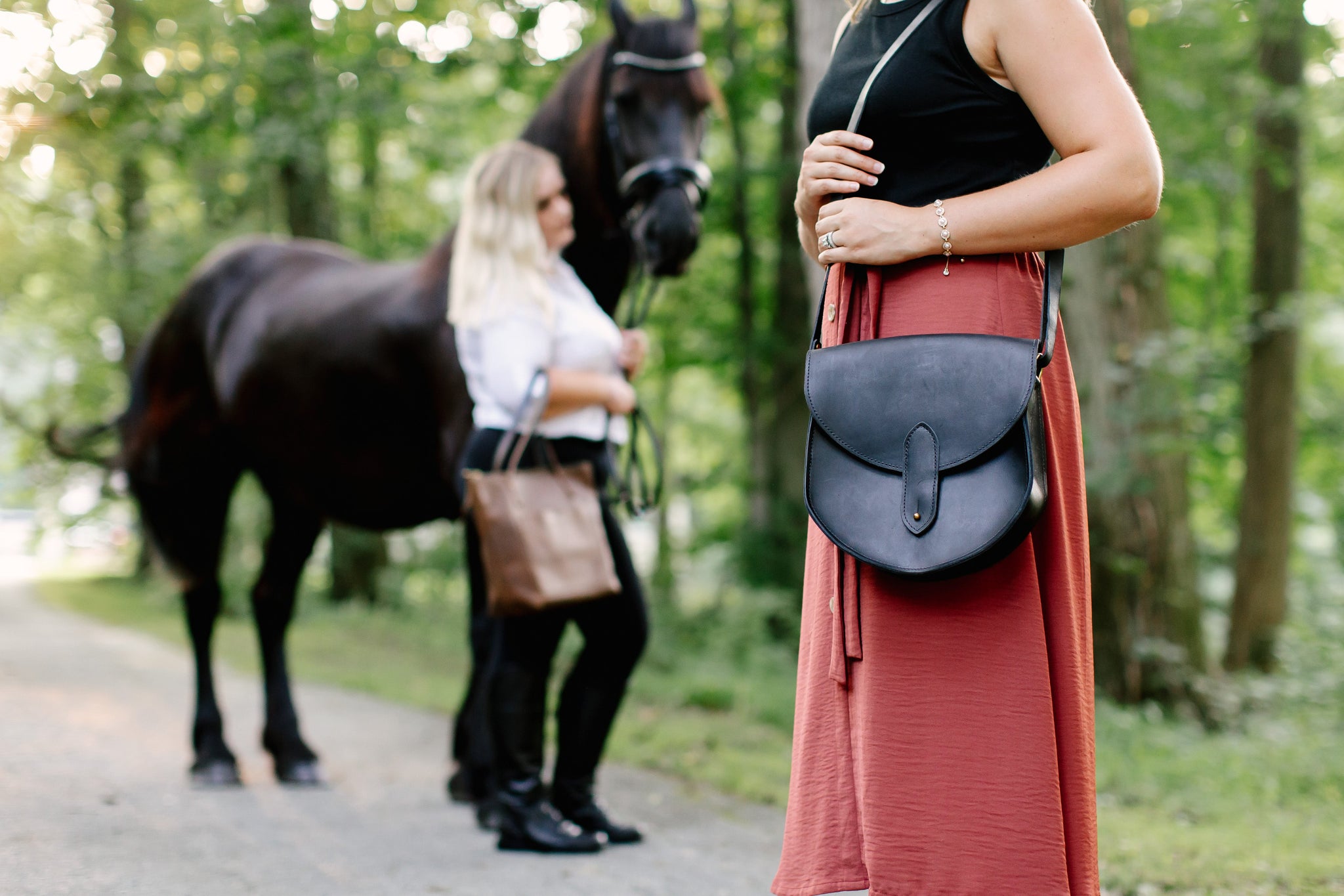 Woman poses with her black leather purse and a Friesian horse on a tree-lined path, while another woman holds the horse's lead.
