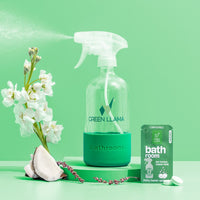 Green Llama bathroom cleaner sprays against a mint green background, showcasing its eco-friendly refill packet, white flowers, and coconut.
