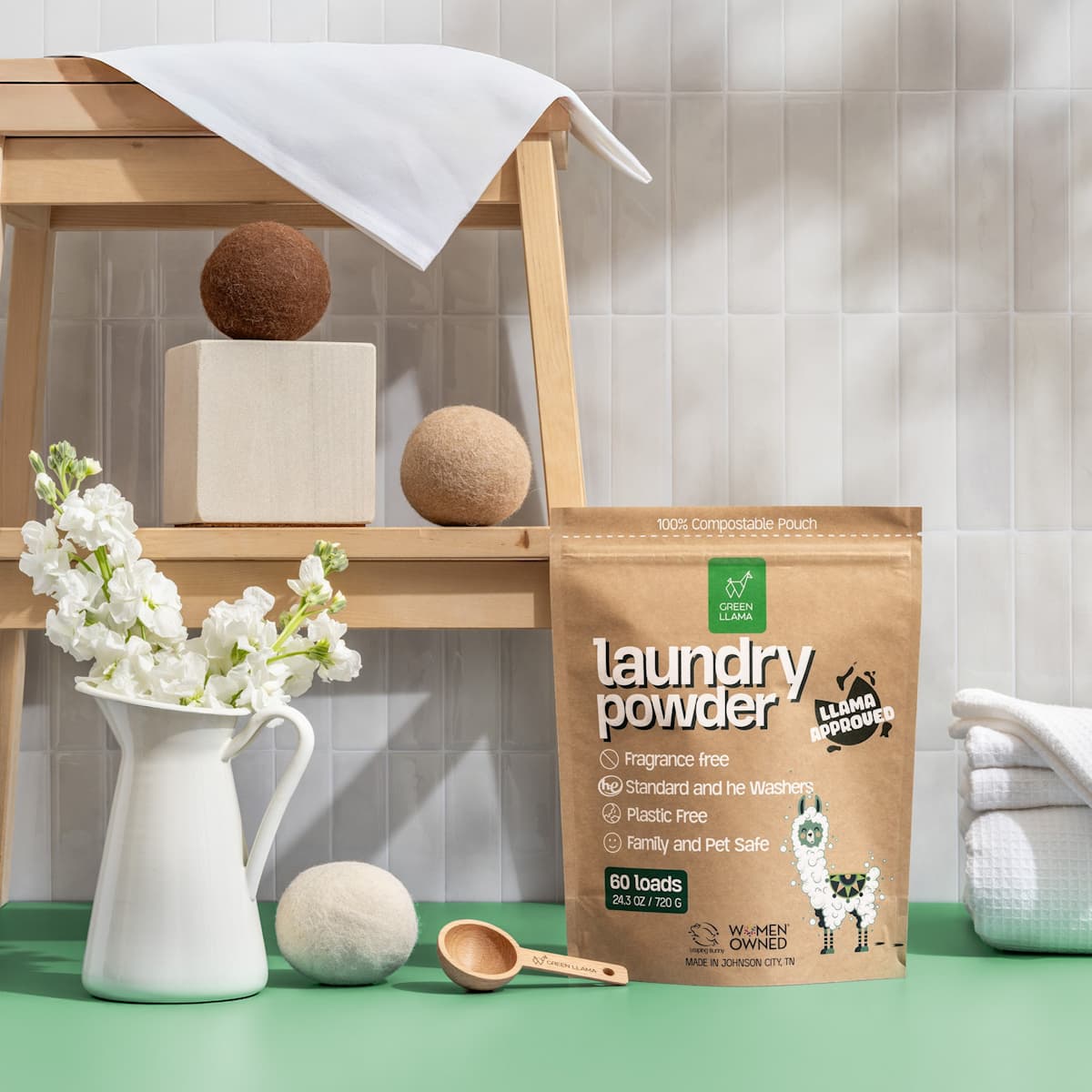 Green Llama laundry powder in a compostable pouch sits near flowers and wool dryer balls in a bathroom setting, showcasing an eco-friendly laundry routine.
