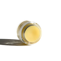 Close-up displays a small glass jar filled with yellow lip balm or salve, casting a soft shadow on a white surface.
