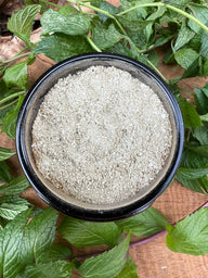 A black bowl contains light gray bath salts, surrounded by fresh mint leaves on a wooden surface.
