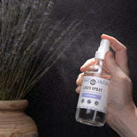 A hand sprays Rustic Strength lavender linen spray near a vase of dried lavender.
