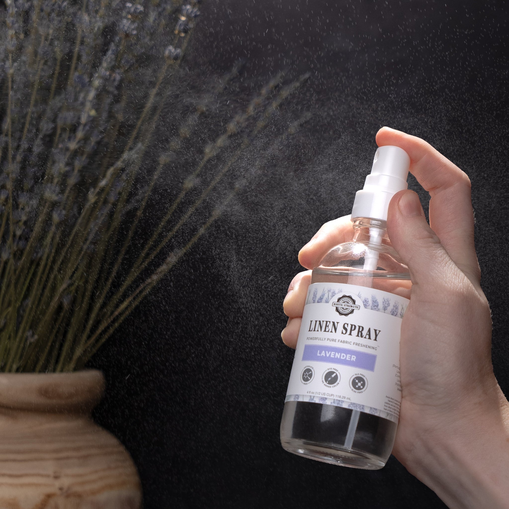 A hand sprays Rustic Strength lavender linen spray near a vase of dried lavender.
