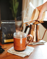 Pouring coffee from a glass filter into a brown leather Urban Southern mug on a wooden table