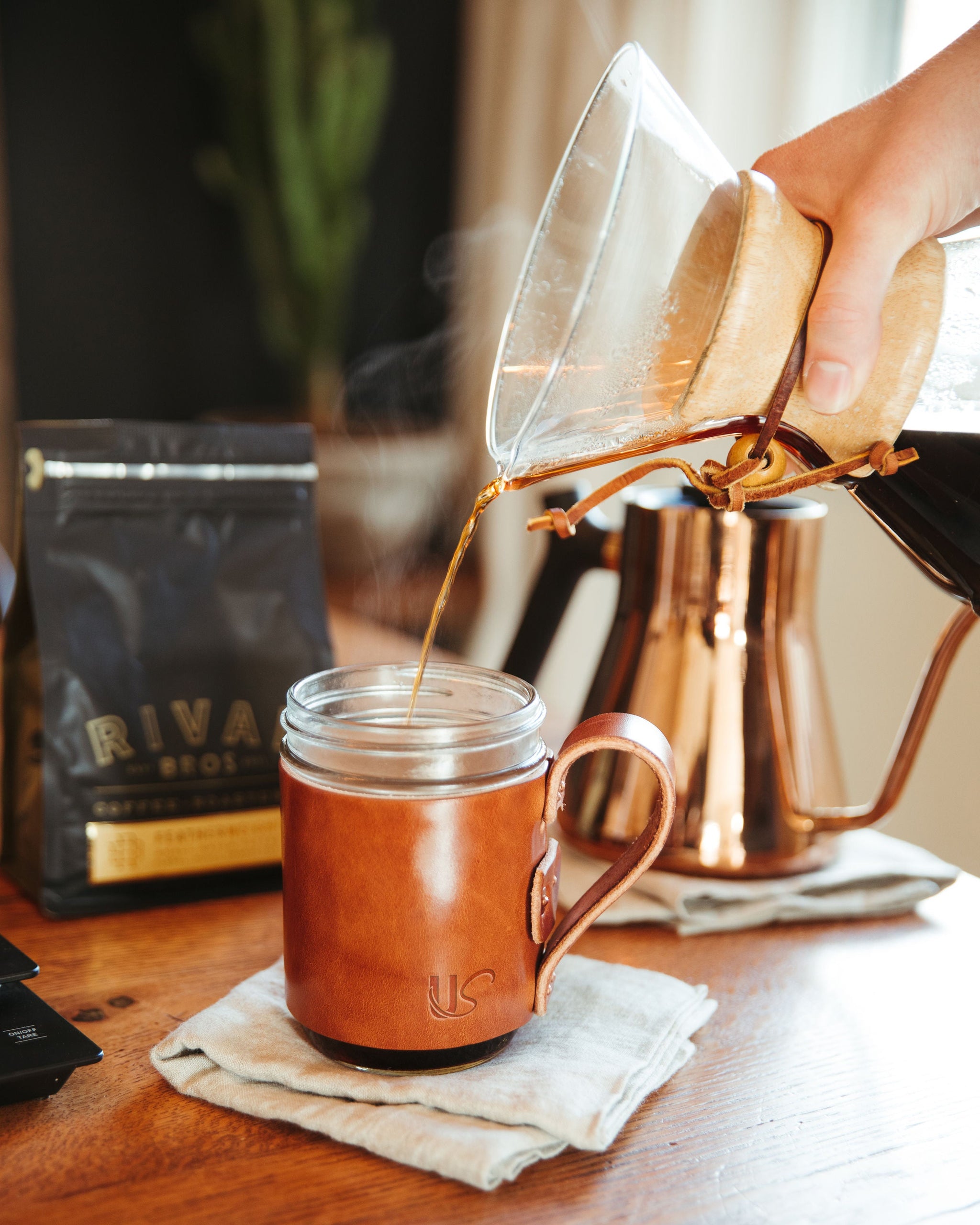 Pouring coffee from a glass filter into a brown leather Urban Southern mug on a wooden table