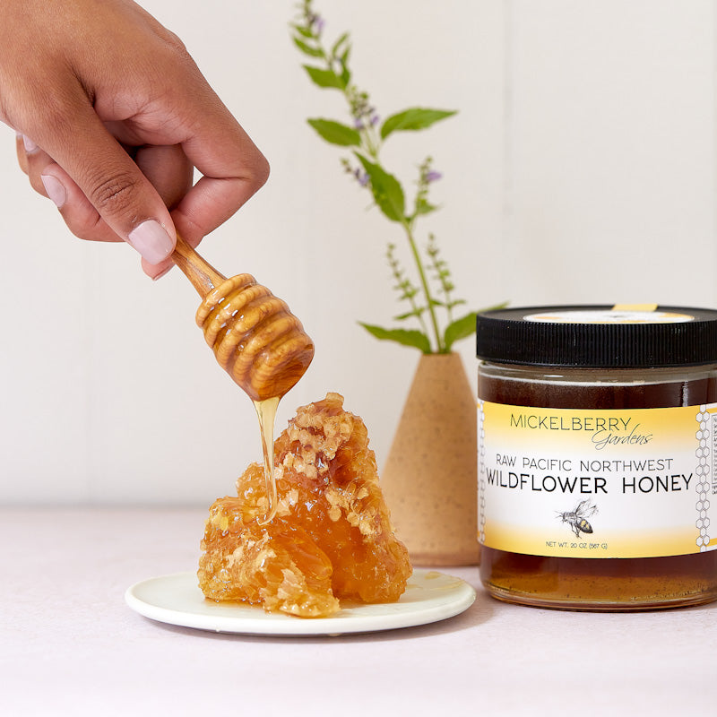 Honeycomb with honey being dripped from a dipper next to a jar of Wildflower Honey on a white surface.