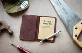 Artisan displays a leather Field Notes journal cover with a notebook and pen alongside a hammer, awl, saw, and snapback hat.
