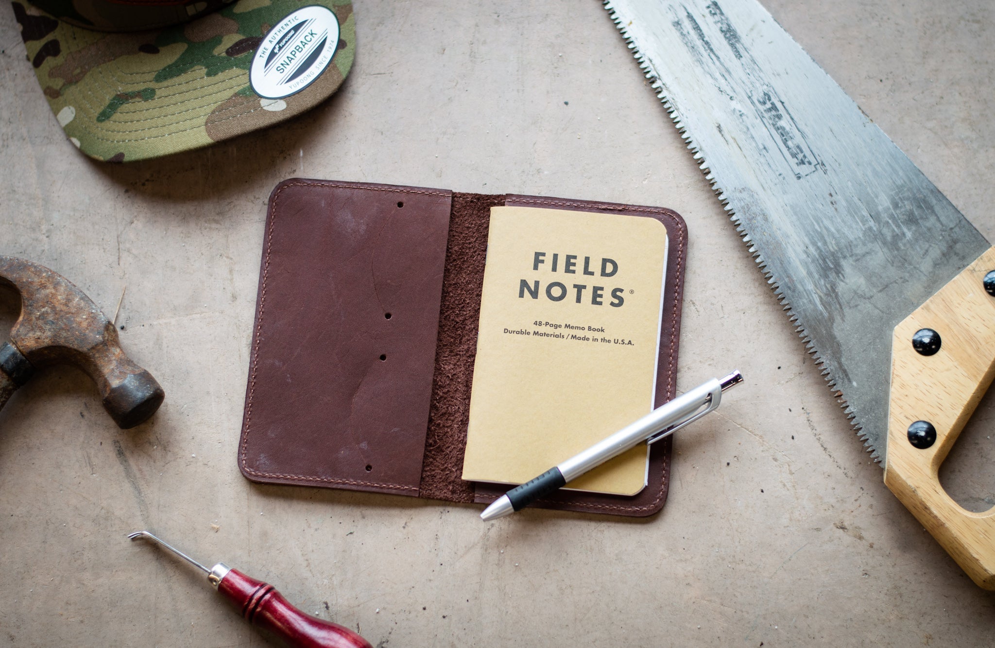 Artisan displays a leather Field Notes journal cover with a notebook and pen alongside a hammer, awl, saw, and snapback hat.
