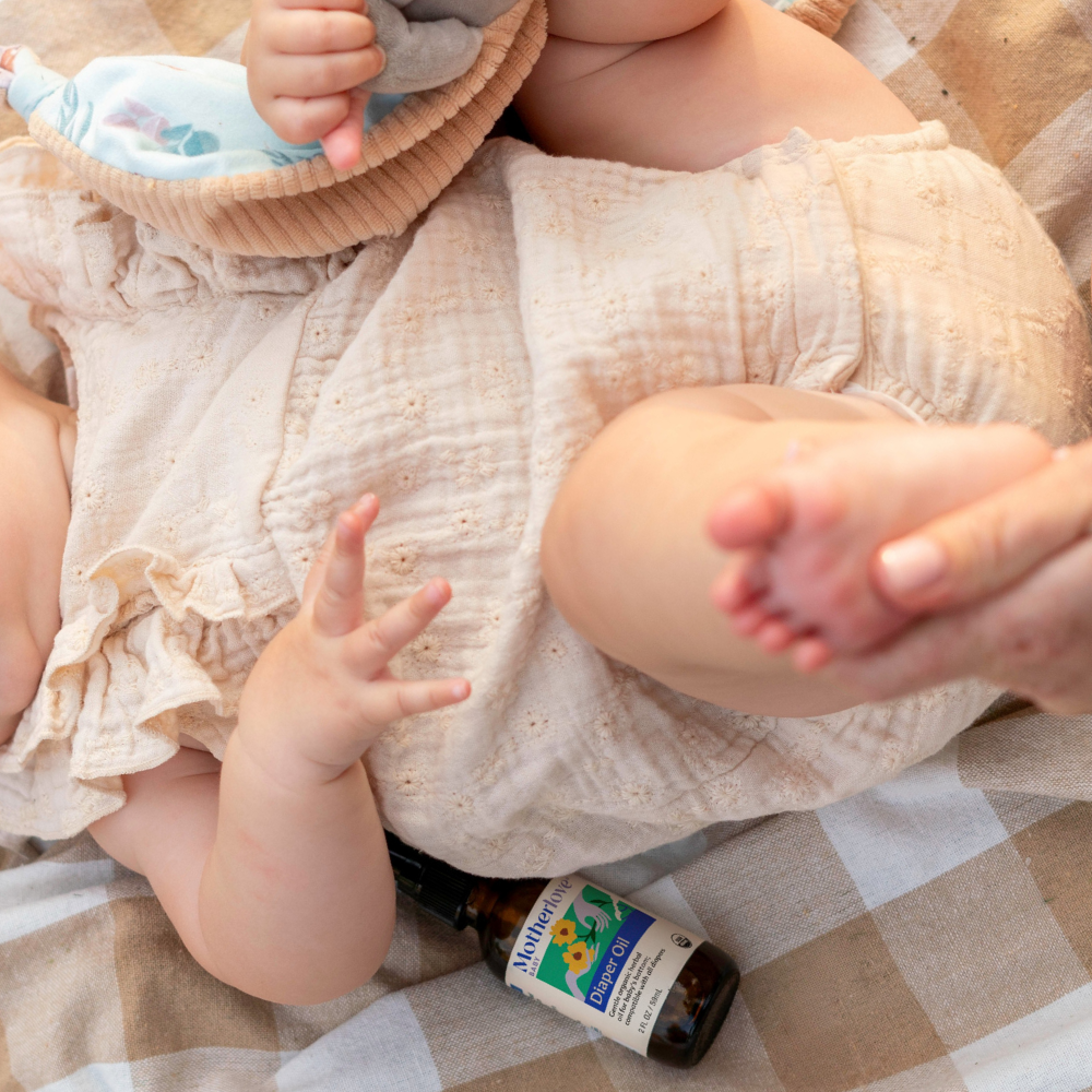 Someone applies Motherlove diaper oil to a baby's foot on a checkered blanket.
