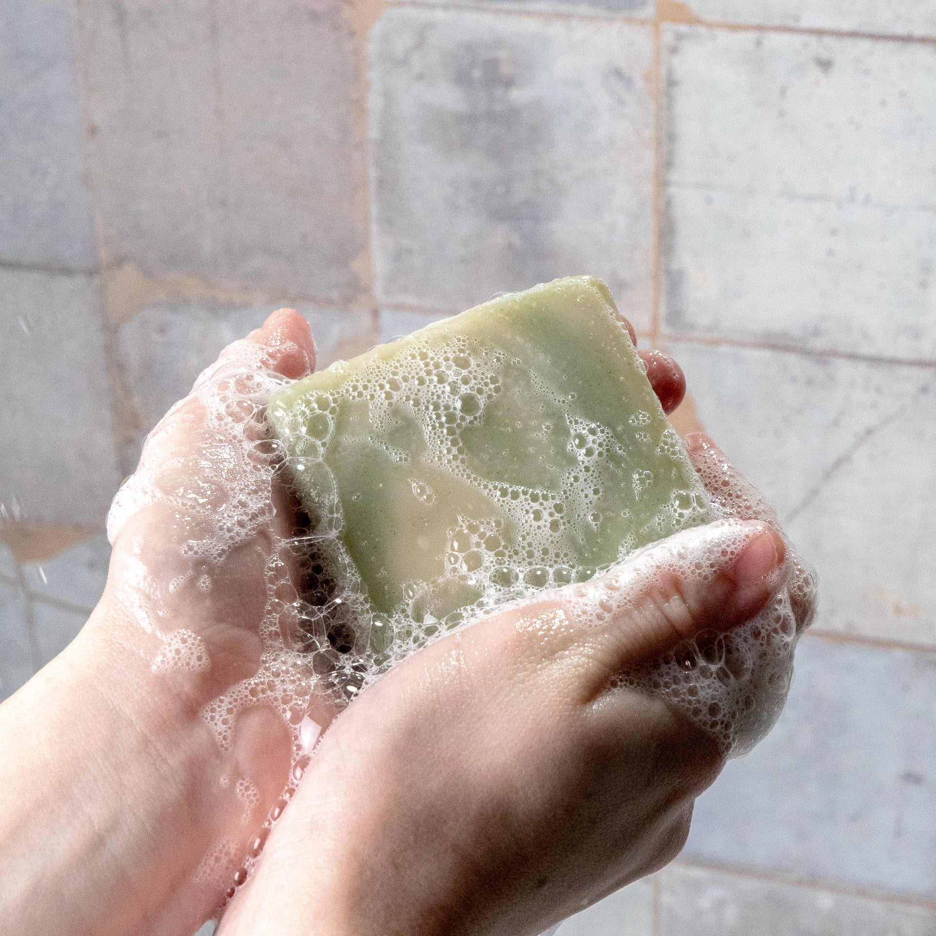Hands lather a bar of natural green soap against a tiled wall.

