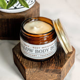 A jar of tallow body butter moisturizer, with mahogany and teakwood scent, sits open on a wooden stand.
