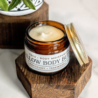 A jar of tallow body butter moisturizer, with mahogany and teakwood scent, sits open on a wooden stand.
