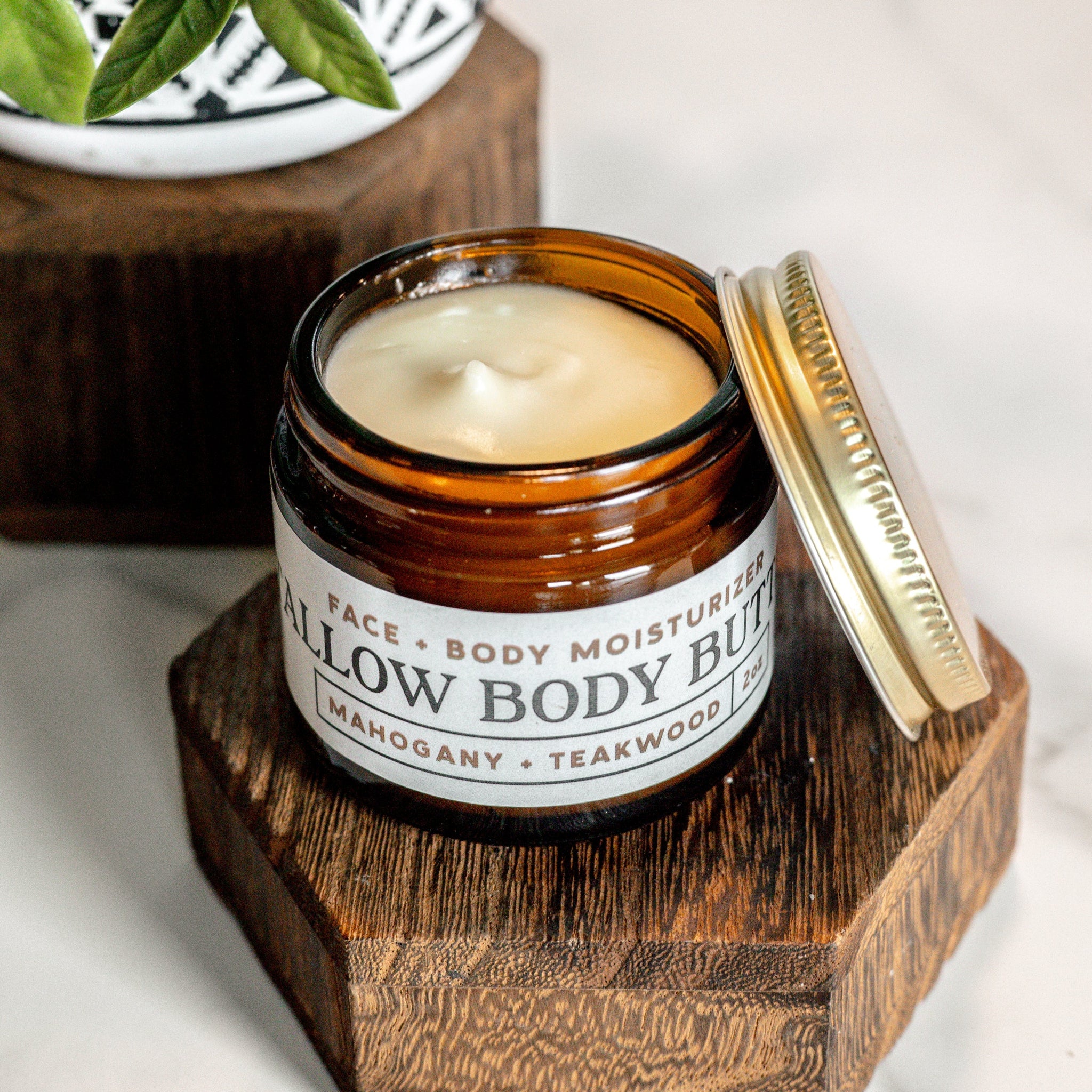 A jar of tallow body butter moisturizer, with mahogany and teakwood scent, sits open on a wooden stand.
