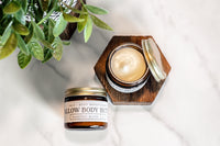 Barefoot Bloom tallow body butter moisturizer sits open on a wooden coaster next to a potted plant.
