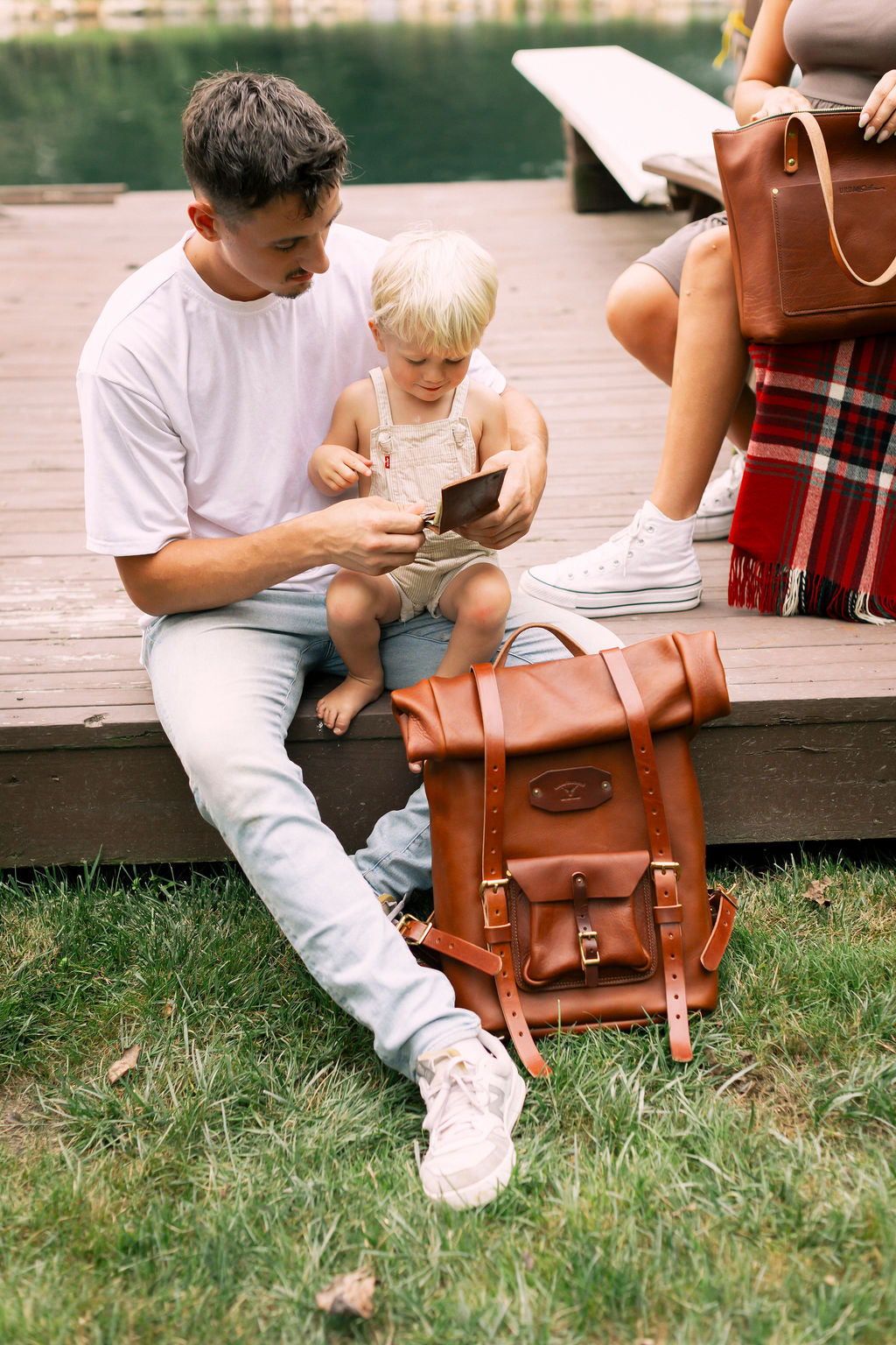 A father shows his young son a leather wallet while sitting on a dock next to a leather backpack and tote bag.
