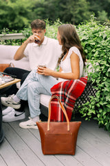 A young couple relaxes on a patio, drinking iced beverages near an Urbano leather tote bag and a red plaid blanket.
