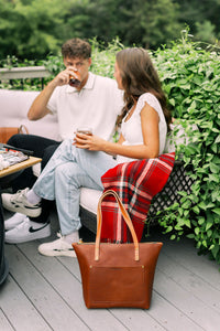 A young couple relaxes on a patio, drinking iced beverages near an Urbano leather tote bag and a red plaid blanket.
