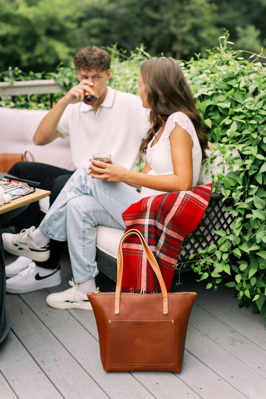 A young couple relaxes on a patio, drinking iced beverages near an Urbano leather tote bag and a red plaid blanket.
