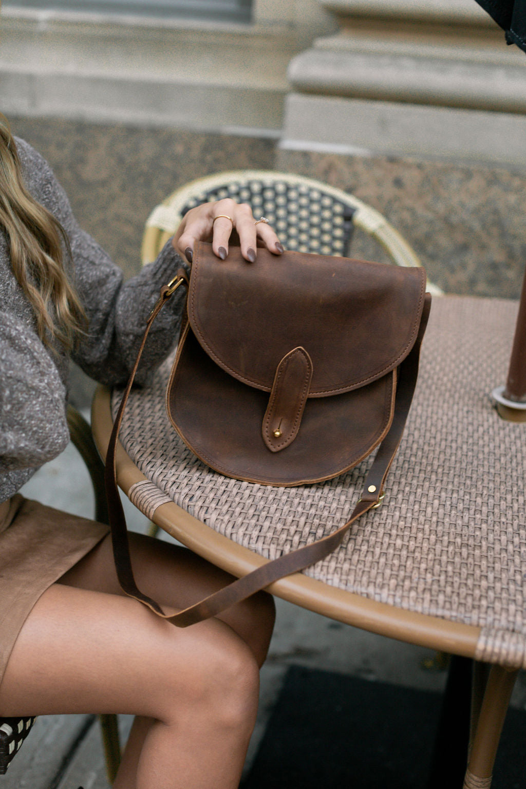 Woman rests her hand on a vintage-style brown leather saddlebag purse on a Parisian cafe table.
